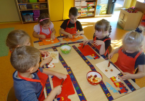 Sześcioro dzieci siedzi przy stole nakrytym podkładkami z rozłożonymi deskami, w ręku trzymają noże, którymi kroją owoce.